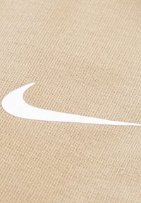 Tissu texturé beige avec un logo Nike Swoosh blanc proéminent. Le matériau est tissé, créant une apparence côtelée subtile.
