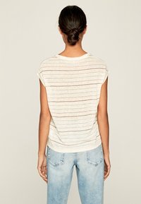 T-shirt z bawełnianego materiału w kolorze białym z cienkimi, poziomymi czerwonymi paskami. Luźny krój, krótki rękaw i okrągły dekolt. Noszony z jasnoniebieskimi dżinsami.