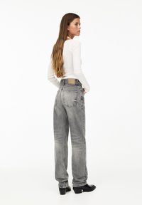 High-waisted, loose-fit grey jeans with a faded texture, featuring back pockets and a tan brand patch on the waistband. Black heeled boots.
