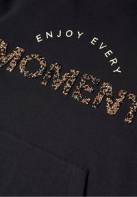 Sweat-shirt noir avec l'inscription "ENJOY EVERY MOMENT" en sequins métalliques texturés. Il présente une texture de tissu lisse et une poche avant.