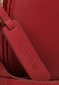 Borsa a tracolla in pelle rossa con texture granulata e tracolla liscia. Etichetta impressa con i dettagli "PICARD" e "SINE 1922".