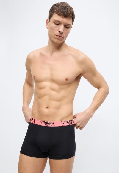 Άνδρας χωρίς πουκάμισο που ρυθμίζει μαύρα σλιπ μποξέρ με ροζ ζώνη μέσης που φέρει το λογότυπο EA σε απλό λευκό φόντο.