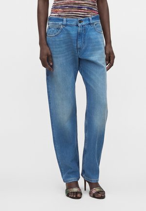 Mørkhudet person iført løstsiddende lyseblå jeans, en flerfarvet stribet top og åbne højhælede sandaler med skinnende stropper.