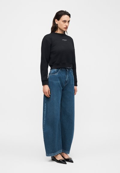 Γυναίκα με μακριά σκούρα μαλλιά, φορώντας μαύρο φούτερ, φαρδιά μπλε τζιν παντελόνια και μαύρα μυτερά φλατ, στέκεται μπροστά σε απλό λευκό φόντο.