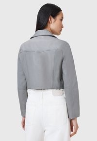 Apgriezta pelēkas krāsas ādas jaka ar platām apkaklēm, gludu tekstūru un šuvju detaļām aizmugurē. Valkāta virs baltām biksēm ar redzamām kabatām.