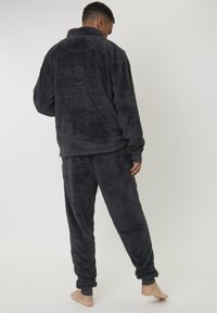 Homme debout pieds nus, portant une veste en polaire fluffy gris foncé et un pantalon assorti, vu de dos sur fond uni.