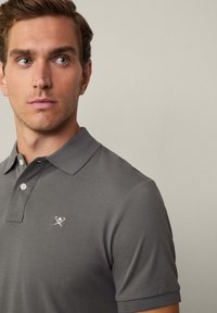 Siva polo majica izrađena od teksturiranog materijala, s ovratnikom, tri dugmeta i malim izvezenim logotipom na lijevoj strani prsnog koša.