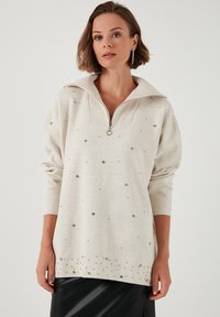 Beżowy sweter z zamkiem przy kołnierzu, ozdobiony srebrnymi detalami z przodu oraz na dole. Wykonany z miękkiego, teksturowanego materiału.