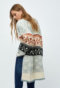 Cardigan en maille crème, bleu, noir et rouille avec des motifs géométriques et symétriques. Comprend des manches longues et une finition douce et texturée.