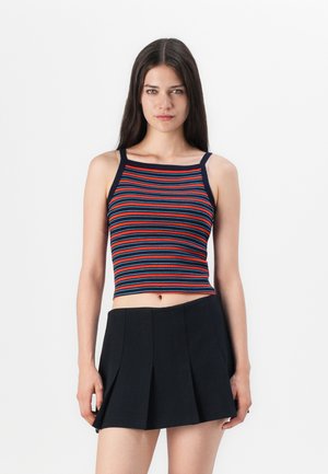 Young woman wearing a sleeveless, striped crop top with navy edges and a black pleated mini skirt, standing against a plain white background.