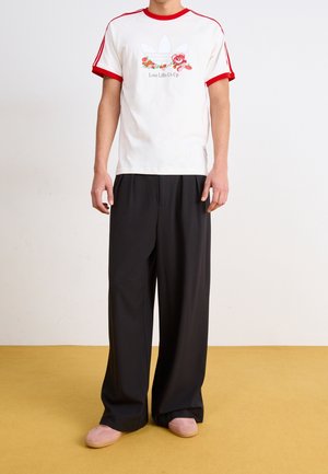 Bílý tričko s červenými akcenty a květinovou grafikou s nápisem "Láska nás pozvedá", v kombinaci s černými širokými kalhotami a růžovými nazouvacími botami.