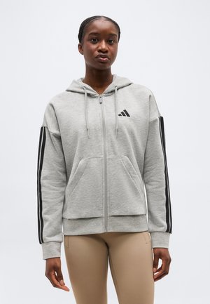 Γυναίκα που φοράει ανοιχτό γκρι φούτερ με φερμουάρ, με μαύρες ρίγες στα μανίκια και λογότυπο, συνδυασμένο με μπεζ εφαρμοστά παντελόνια, στέκεται μπροστά σε λευκό φόντο.