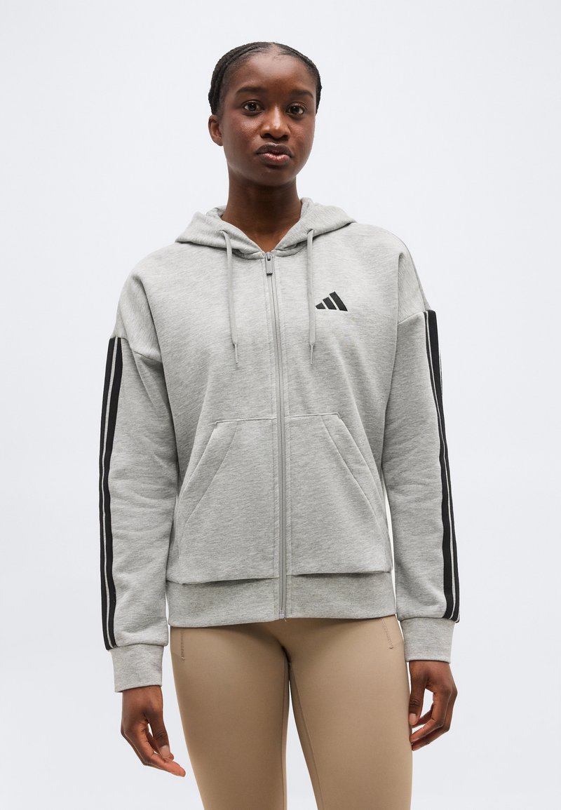 Sieviete valkā gaiši pelēku rāvējslēdzēja kapuci ar melnām svītrām uz piedurknēm un logotipu, kopā ar bēša krāsas piegulošām biksēm, stāvot pret baltu fonu.