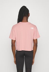 Korte mouwen cropped t-shirt in lichtroze katoen, met een klein wit logo op de achterkant nabij de halslijn. Gedragen met zwarte leggings.