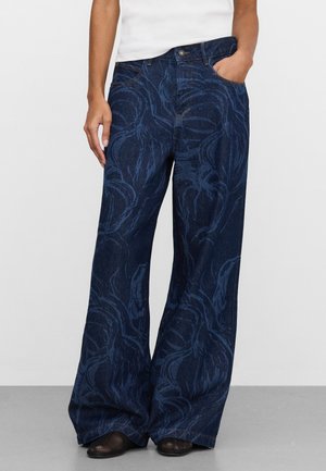 Osoba ubrana w ciemnoniebieskie, szerokie dżinsy z delikatnym kwiatowym wzorem, biały top i czarne buty, stojąca z jedną ręką w kieszeni.