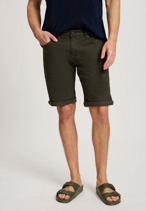 Olivgrüne Baumwollshorts mit umgeschlagenen Säumen, Fronttaschen und einem Knopfverschluss, kombiniert mit dunkelgrünen Gummisandalen.