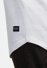 Biała koszulka z bawełny o prostym kroju. Ma naszytą czarną metkę Jacks & Jones na rękawie. Czarne dżinsy są częściowo widoczne.