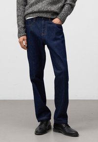 Ciemnoniebieskie dżinsy o prostym kroju, z widocznymi kieszeniami i kontrastującymi pomarańczowymi przeszyciami. Ubrane z teksturowanym szarym swetrem i czarnymi butami.