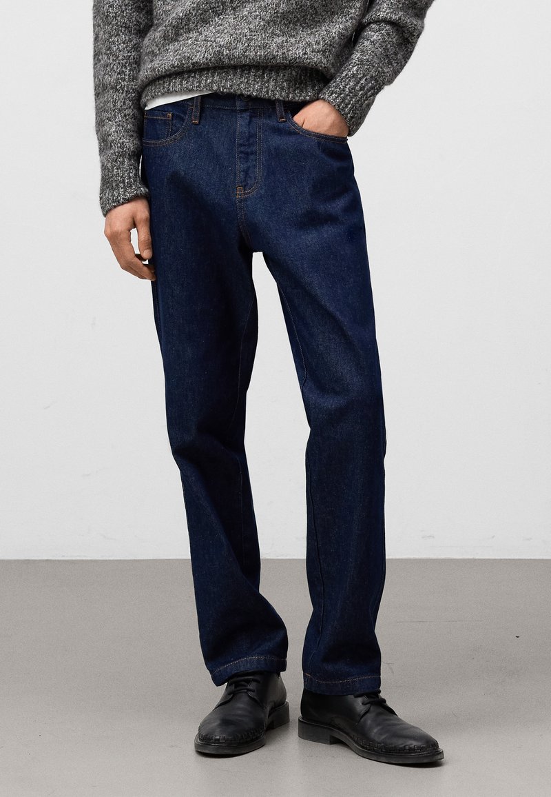 Ciemnoniebieskie dżinsy o prostym kroju, z widocznymi kieszeniami i kontrastującymi pomarańczowymi przeszyciami. Ubrane z teksturowanym szarym swetrem i czarnymi butami.