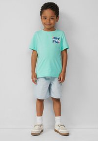 Turquoise T-shirt met korte mouwen met "GET FUN" grafiek, gecombineerd met lichtblauwe shorts en witte sokken, gedragen met crèmekleurige sneakers.
