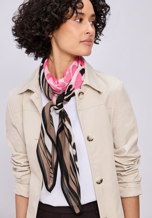 Donna con capelli ricci che indossa una giacca beige e una camicia bianca, guarda verso destra, sciarpa rosa e marrone con motivo annodata intorno al collo.