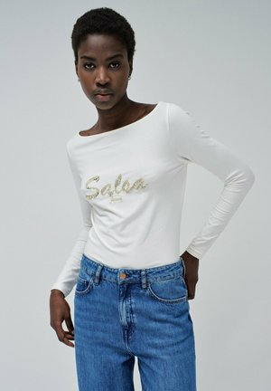 Top blanco de manga larga con el texto "Salsa" en acentos brillantes, combinado con jeans de mezclilla azul de talle alto. Diseño simple y ajustado.
