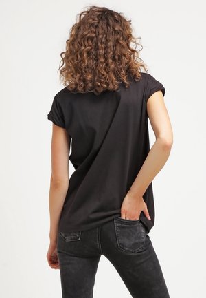 Kobieta z kręconymi włosami, ubrana w czarny t-shirt z podwiniętymi rękawami i ciemnoszare jeansy, z jedną ręką w tylnej kieszeni, zwrócona plecami.