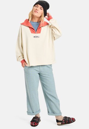 Helle, übergroße Sweatshirt mit einem korallenfarbenen Kragen und Logo, kombiniert mit lockeren grünen Karo-Hosen und schwarzen Rutsch-Sandalen.