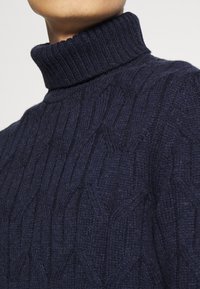 Gros plan sur un pull à col roulé bleu marine en maille torsadée porté par une personne, montrant le col côtelé texturé et le motif en tricot.