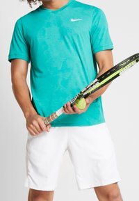 Person som bär en turkos Nike-tröja och vita shorts och håller en tennisracket och en boll mot vit bakgrund.