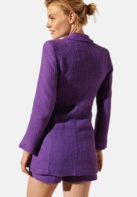 Blazer texturé violet avec des manches longues, un col et une coupe ajustée, associé à un short assorti, mettant en valeur un design en tissu tissé.