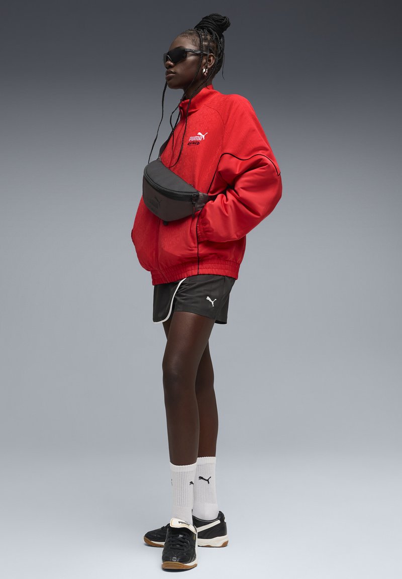Veste bomber rouge avec accents noirs, sac banane noir, short de sport noir, chaussettes blanches et baskets noires. Des lunettes de soleil complètent le look.