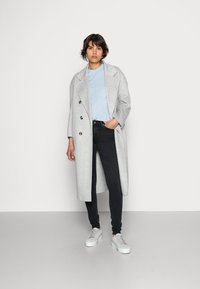 Casaco cinza claro, de botões duplos e grandes, usado sobre uma t-shirt azul e jeans skinny pretos; combinado com ténis cinza claro.