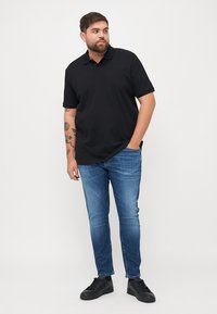 Fekete, rövid ujjú póló, sima felülettel, kék, slim-fit farmerrel és fekete sportcipővel párosítva, egy sima háttér előtt állva.