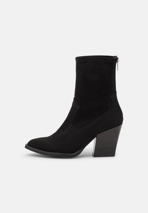 Bottine en suede noir avec un bout pointu, des panneaux élastiques sur les côtés et un talon bloc en bois épais. Elle dispose d'une fermeture éclair à l'arrière.