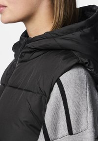 Svart vadderad väst med huva, med quiltad textur och hög krage, lagd över en ljusgrå tröja med svarta ränder.