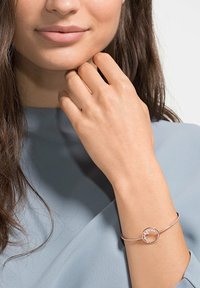 Roségold-Armband mit einem runden Anhänger, der ein kleines Edelstein-Detail aufweist, getragen an einer Hand vor einem weichen blauen Kleidungsstück. Glatte metallische Oberfläche.