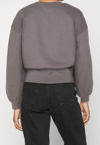 Sweatshirt grigio con una vestibilità rilassata, maniche a palloncino e dettagli dorati sparsi. Presenta orli e polsini a coste. Indossato con pantaloni neri.