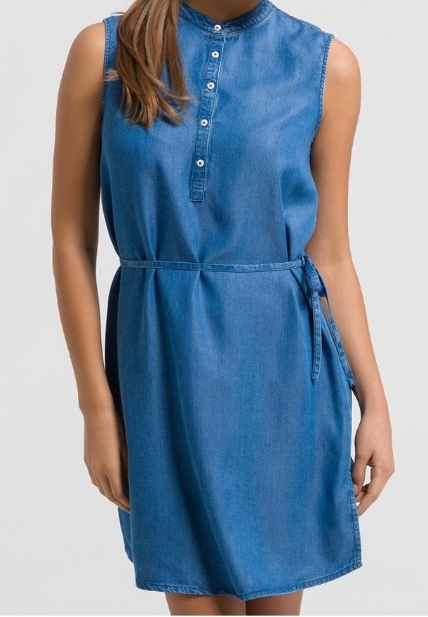 Robe en jean bleu sans manches avec un col boutonné et une fine ceinture nouée à la taille, portée par une personne aux cheveux longs.