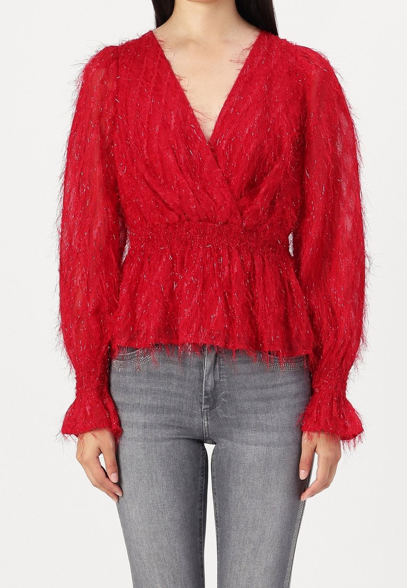 Blouse rouge texturée à manches longues avec col en V et taille froncée portée avec un jean gris clair sur une personne debout.