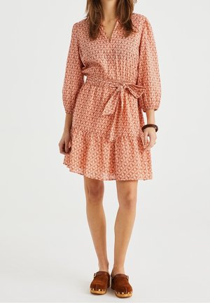 Růžové šaty se vzorem, V-krkem, balonovými rukávy a páskem v pase, s řaseným lemem. Kombinovány s hnědými dřeváky pro kontrast.