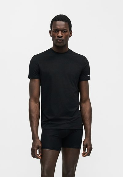 Άνδρας με εφαρμοστή μαύρη κοντομάνικη μπλούζα και μαύρο μπόξερ, στέκεται μπροστά σε απλό ανοιχτό φόντο.