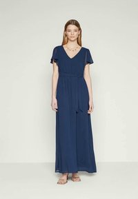 Jovem mulher em pé, vestindo um vestido azul marinho até ao chão, com mangas esvoaçantes, decote em V, cintura amarrada e sandálias rasas, sobre um fundo simples.