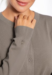 Helle graue Strickjacke aus geripptem Stoff, mit Knopfverschluss und gesticktem Logodetail am Bund. Weicher Stoff und taillierte Form.