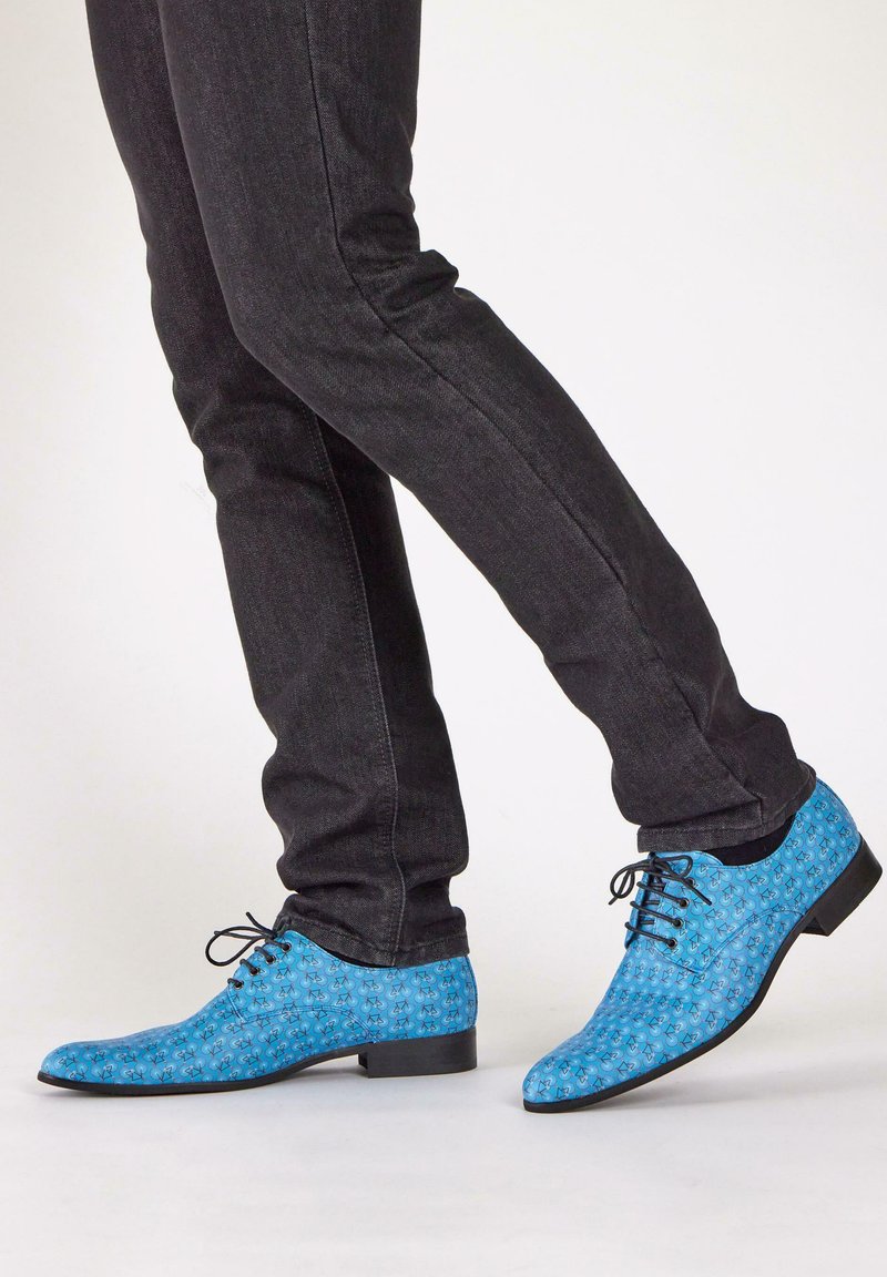 Blauwe schoenen met een gladde textuur, voorzien van een bloemenpatroon. Glanzende zwarte zolen en vetersluiting, gecombineerd met een donker paar jeans.