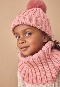 Rosa gestrickte Mütze mit Rippstrickdesign und Pompon oben, kombiniert mit einem passenden Rippstrick-Nackenwärmer. Weicher Stoff vor einem neutralen Hintergrund.