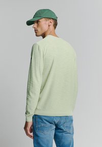 Lichtgroene blouse met lange mouwen en een textuurafwerking, geribde manchetten en zoom, gecombineerd met blauwe denim jeans. Het model draagt een groene pet.
