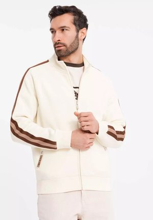 Cream zip-up jacket with brown and beige stripes on sleeves. Features a high collar, ribbed cuffs, and a logo tag at the hem.