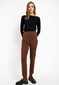 Kobieta stoi z rękami za plecami, ubrana w czarną bluzkę z długim rękawem, brązowe spodnie z wysokim stanem i czarne platformowe lordsy na białym tle.