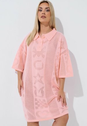 Kobieta o długich blond włosach ubrana w luźną, jasnoróżową dzianinową sukienkę z kołnierzykiem i wzorzystymi geometrycznymi wycięciami.